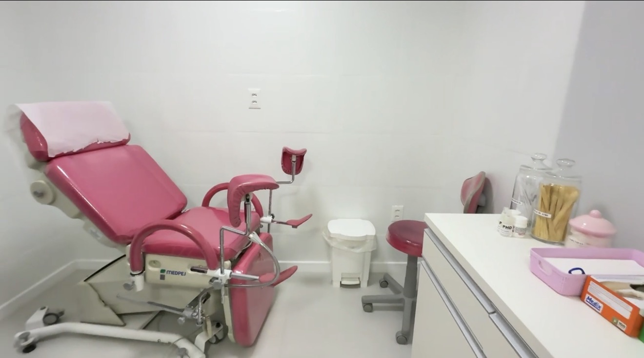 Sala de atendimento equipada com maca cor-de-rosa, banco giratório e bancada com materiais descartáveis